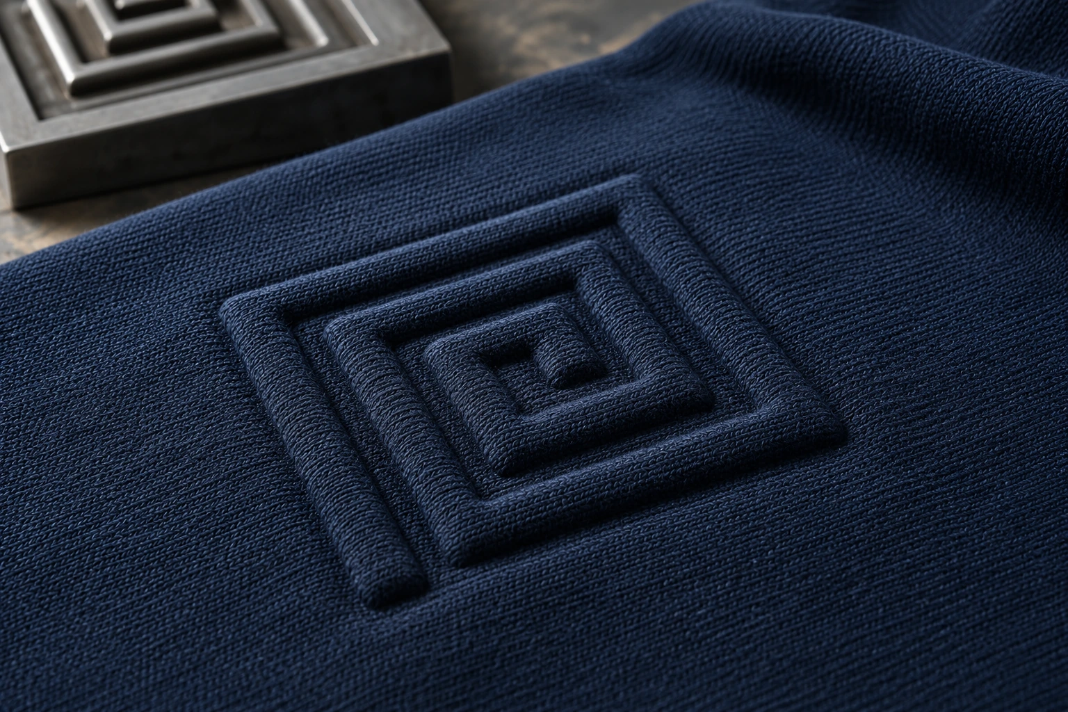 Tonal geometric embossed relief in navy knit fabric beside a matching metal die.